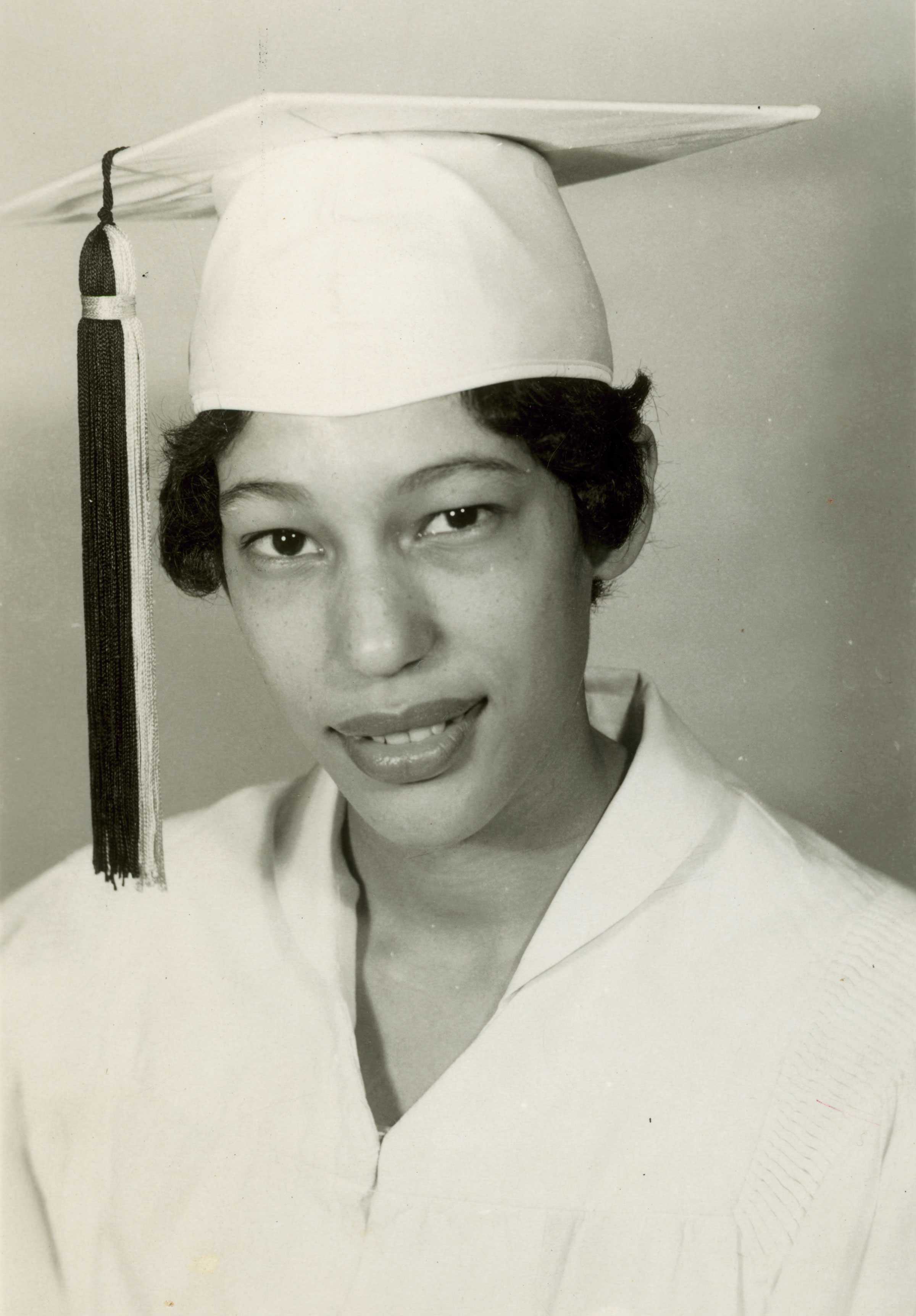 Carlotta Walls LaNier smiles in a white graduation gown and hat for her graduation portrait. She has short wavy hair.