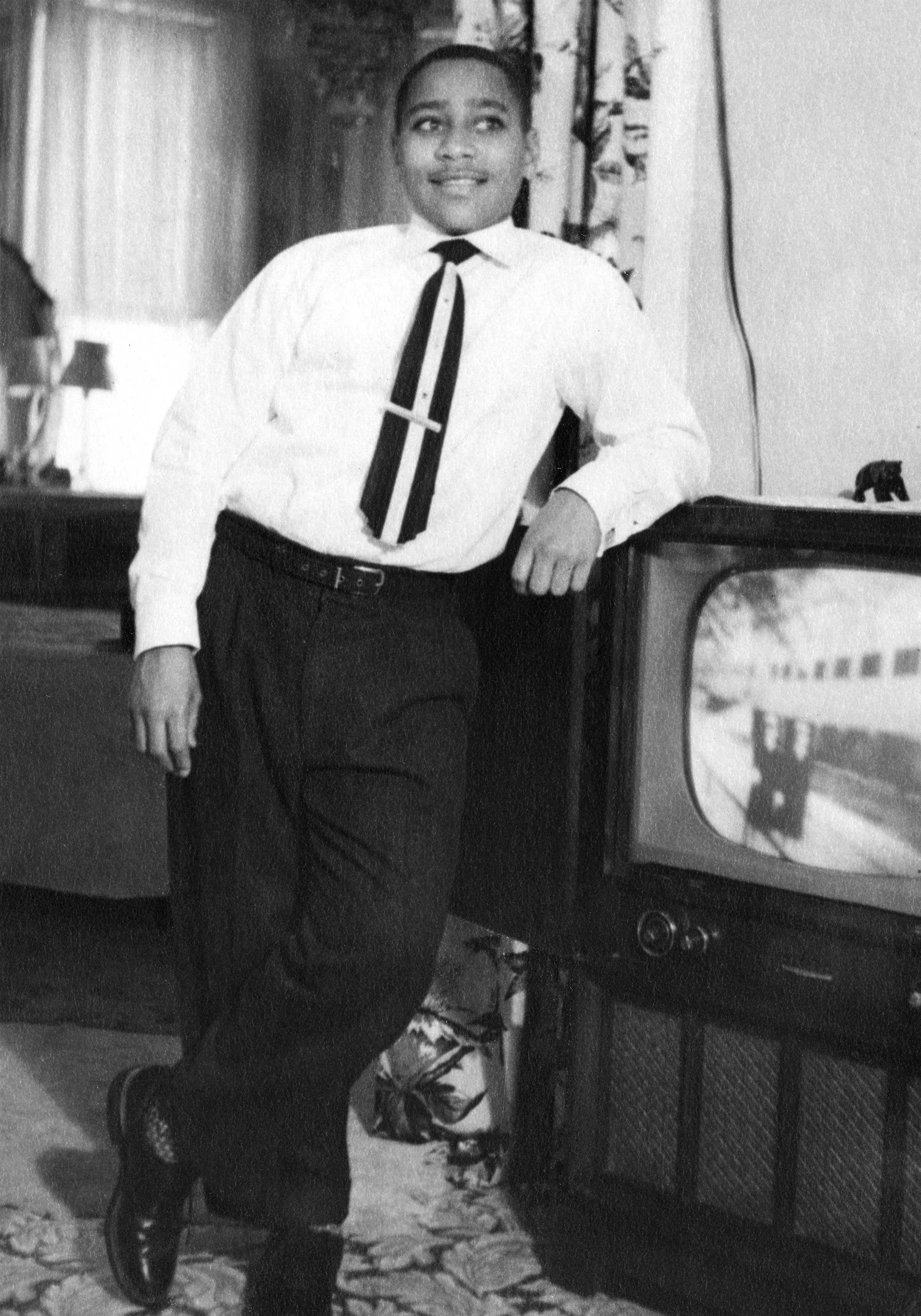 13 years old Emmett Till at thirteen leans on a television set as he smiles for the camera. He is dressed in a white dress shirt and black pants, with a white and black tie.