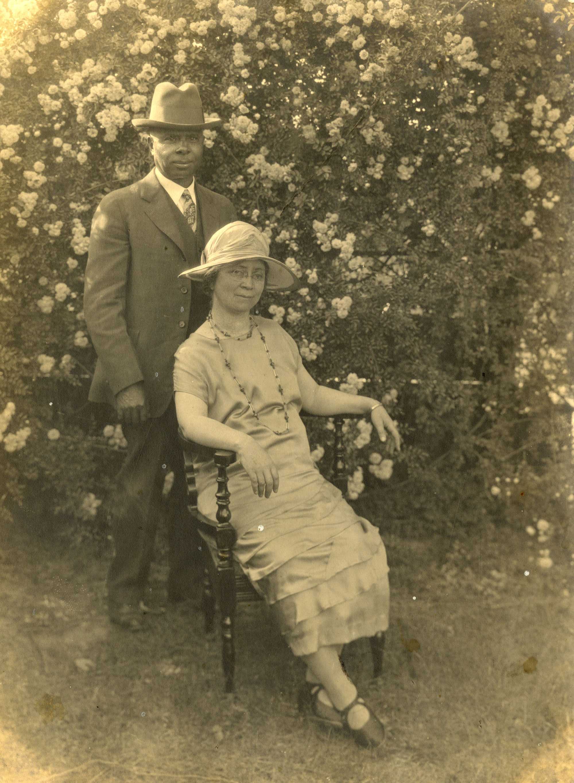 A black and white photo of two people in a garden, one standing and one sitting in a chair.