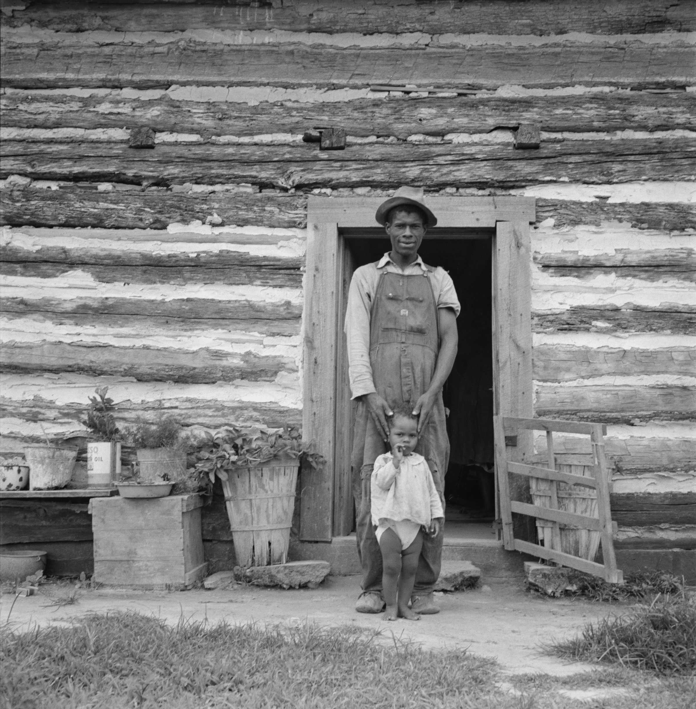 Black and white photograph of an African American man with small child standing outside doorway of wooden house.