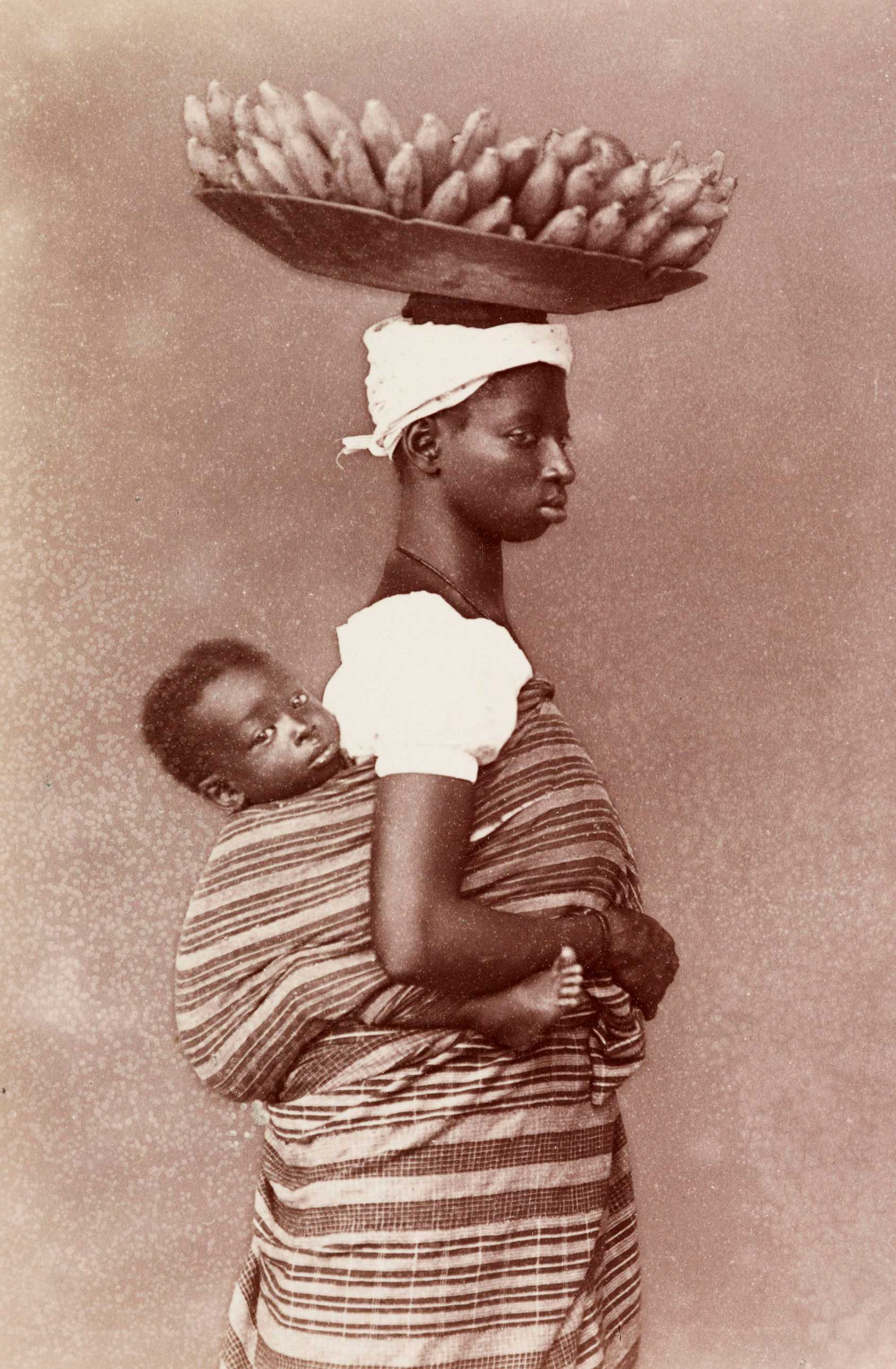 A black and white photograph of an enslaved woman with her child wrapped on her back and a bowl of plantains on her head.