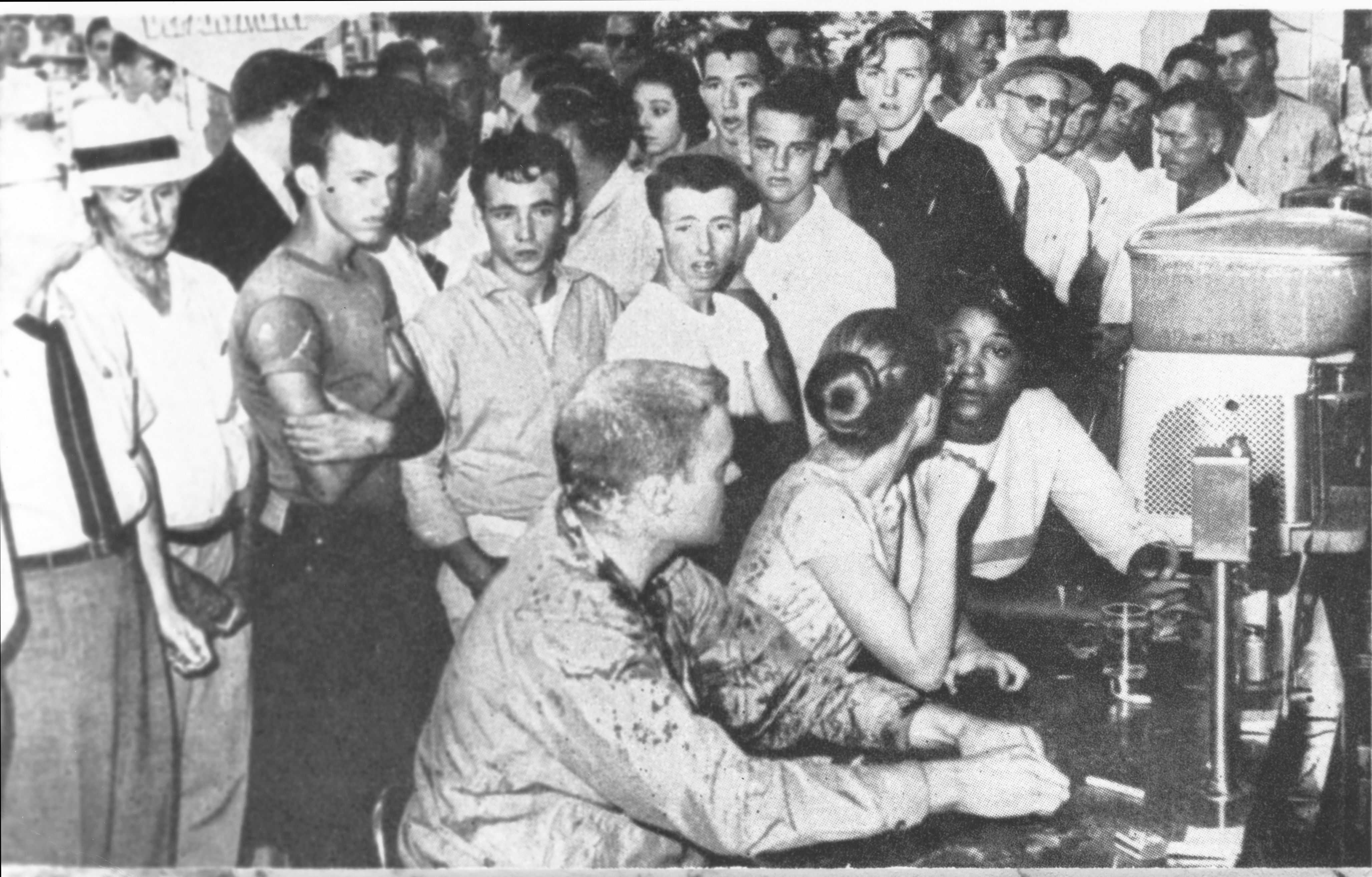 Black and white photograph showing students sitting at lunch counter while a large group of onlookers stand behind them.