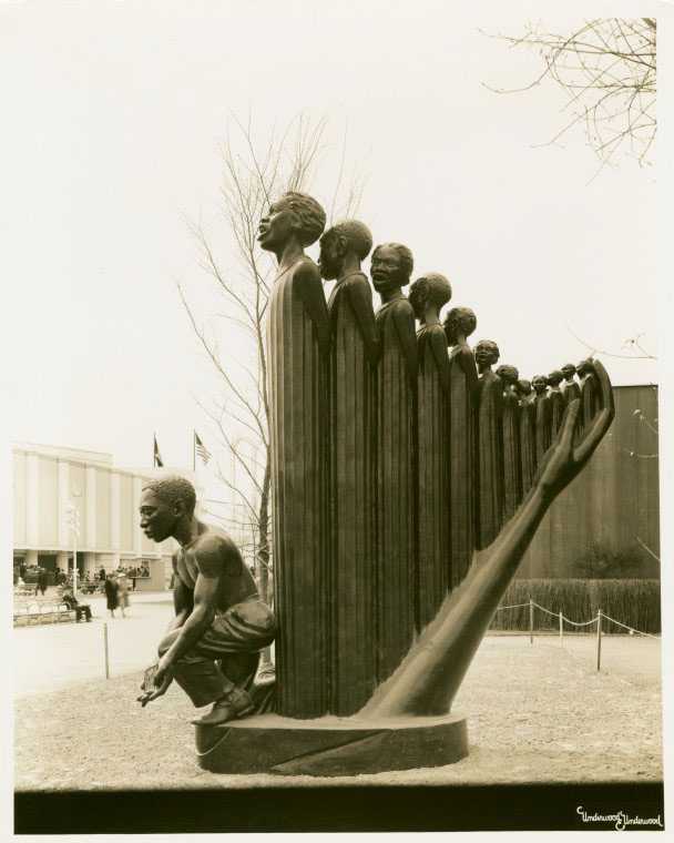 An artistic sculpture composed of multiple human heads, gradually increasing in size from left to right. In front of these heads, a crouched figure extends an arm upwards. he background features leafless trees, an American flag on a flagpole, and part of a building with obscured lettering.