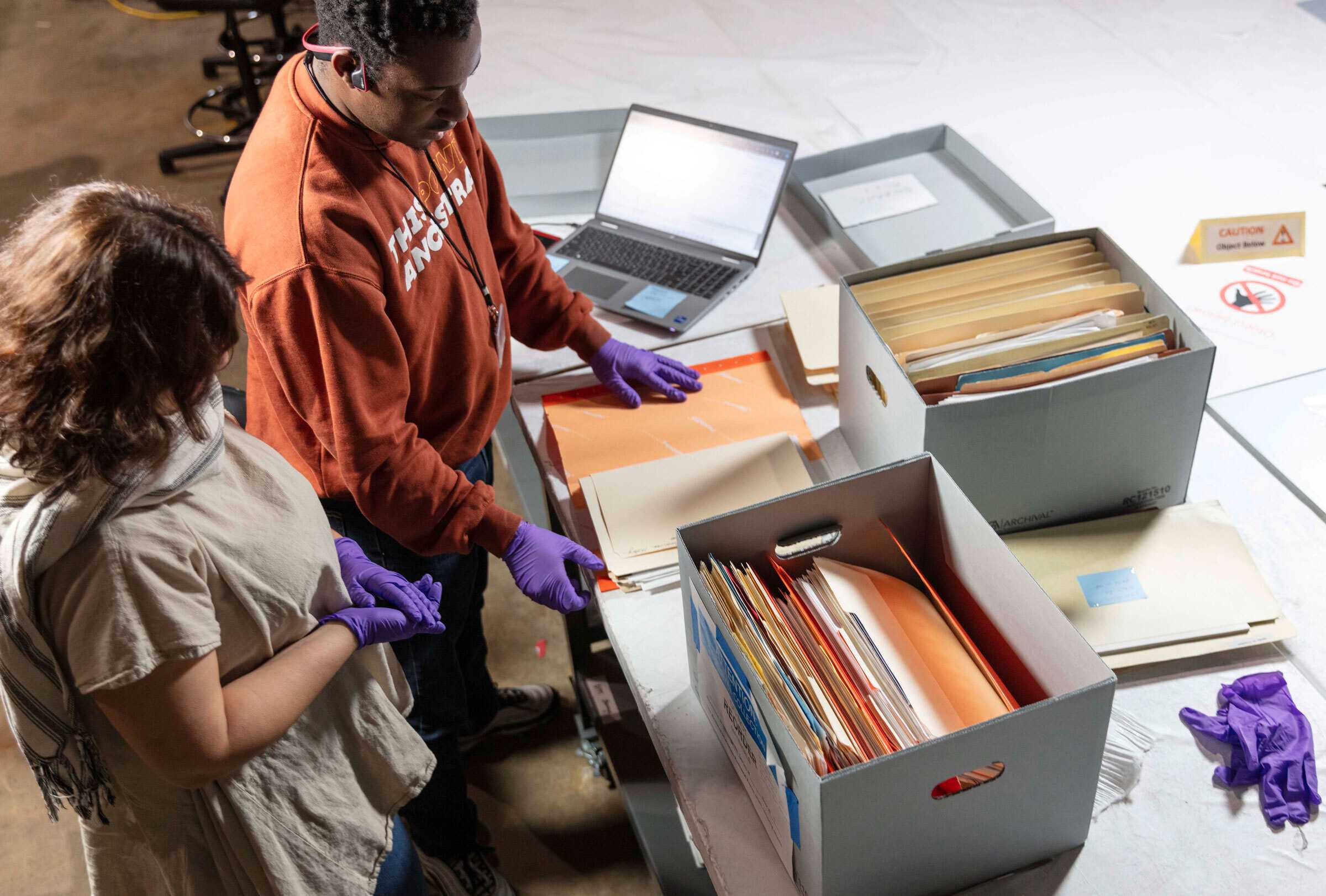 Two people wearing gloves review paper files from archival boxes on a worktable with an open laptop nearby.