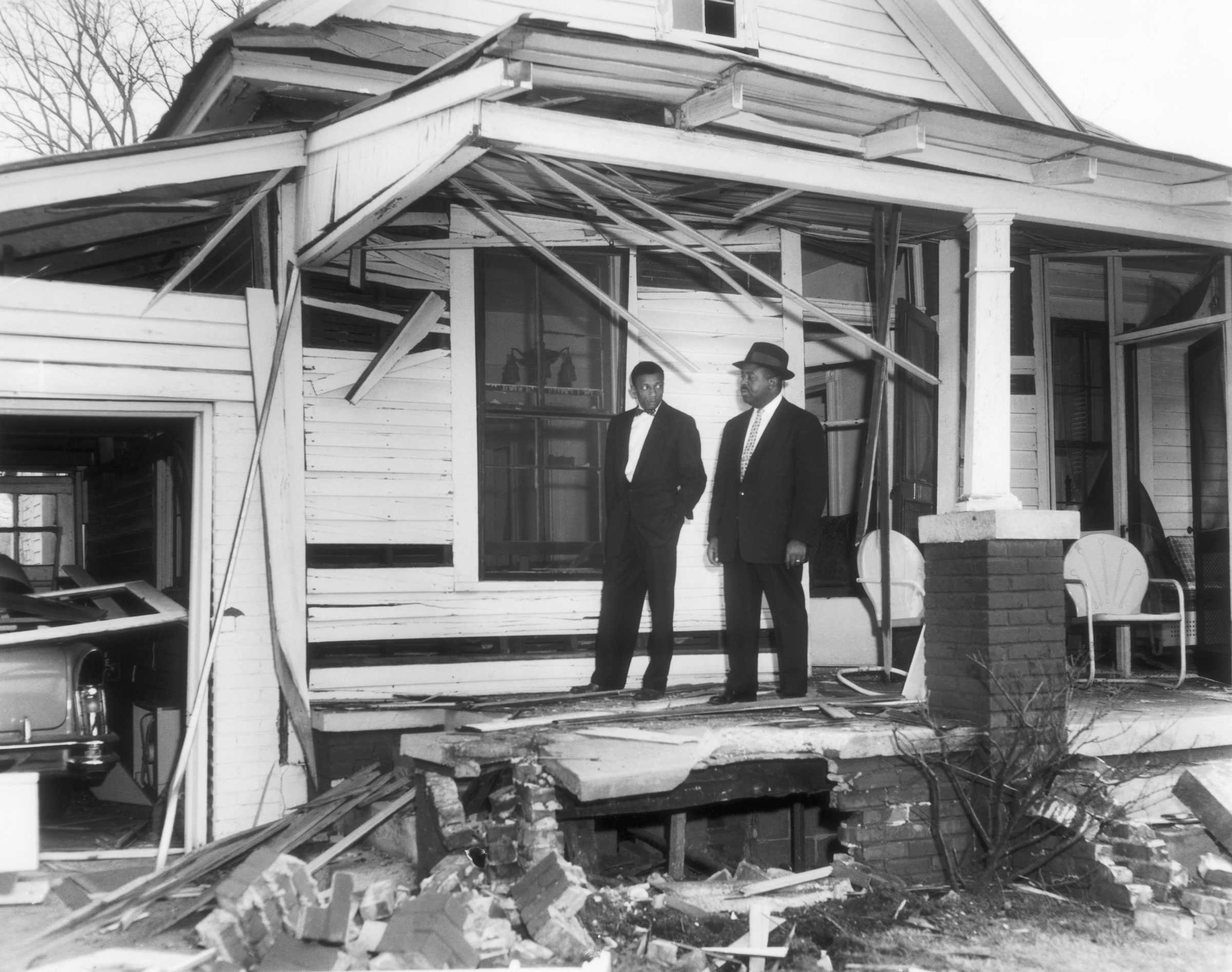 A black and white photograph of Rev. Ralph Abernathy's damaged house. Two men in suits stand on the remain part of the porch.