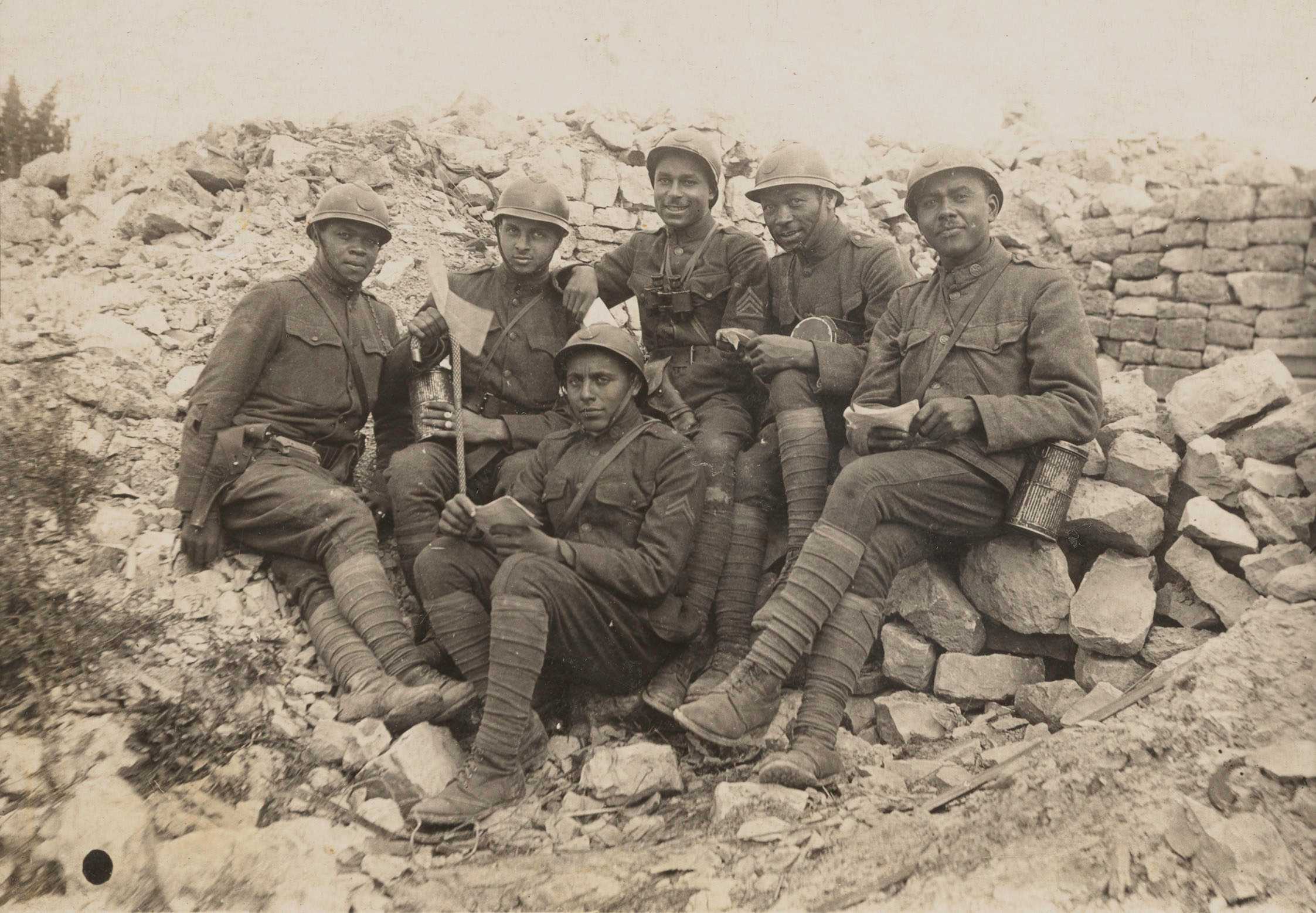 A postcard shows Oscar Calmeise (far right) and five unidentified soldiers in WWI, sitting on stones in Verdun, France. They wear helmets and uniforms.