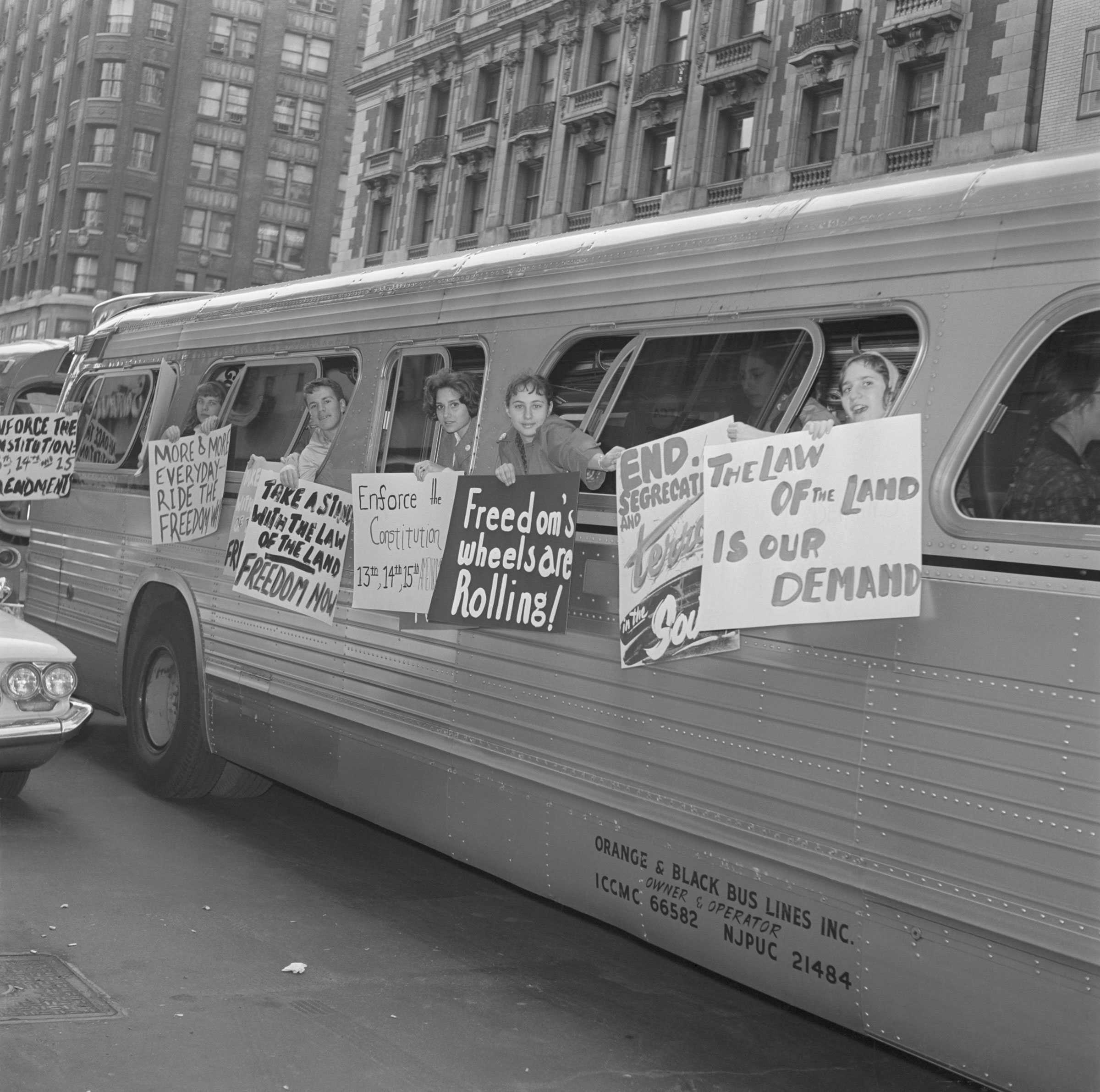 White demonstrators hold signs outside of a Freedom bus, demanding Civil Rights, as they drive on a street.