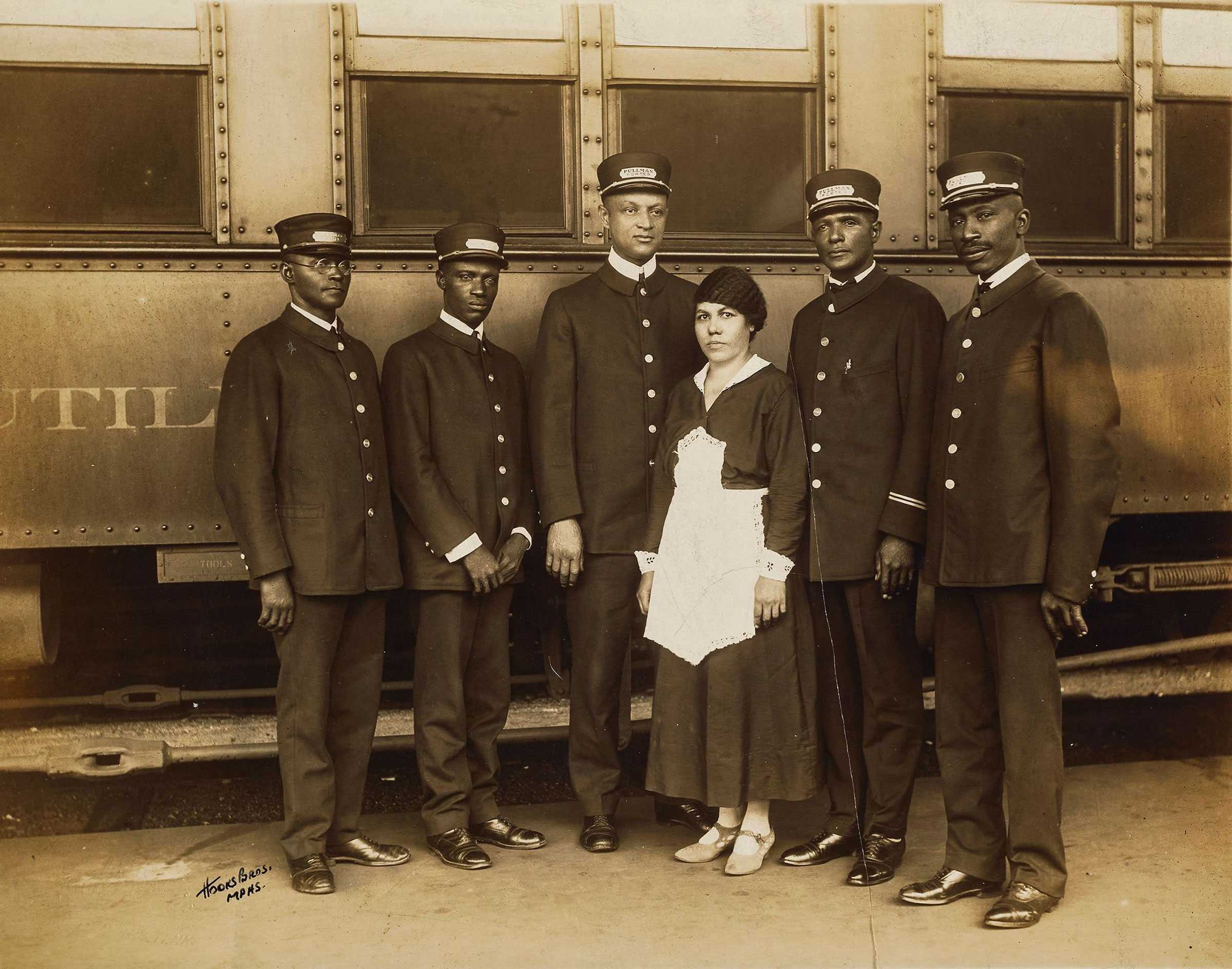 A sepia toned photograph of 5 pullman porters and one maid, posing for a portrait outside of a train car.