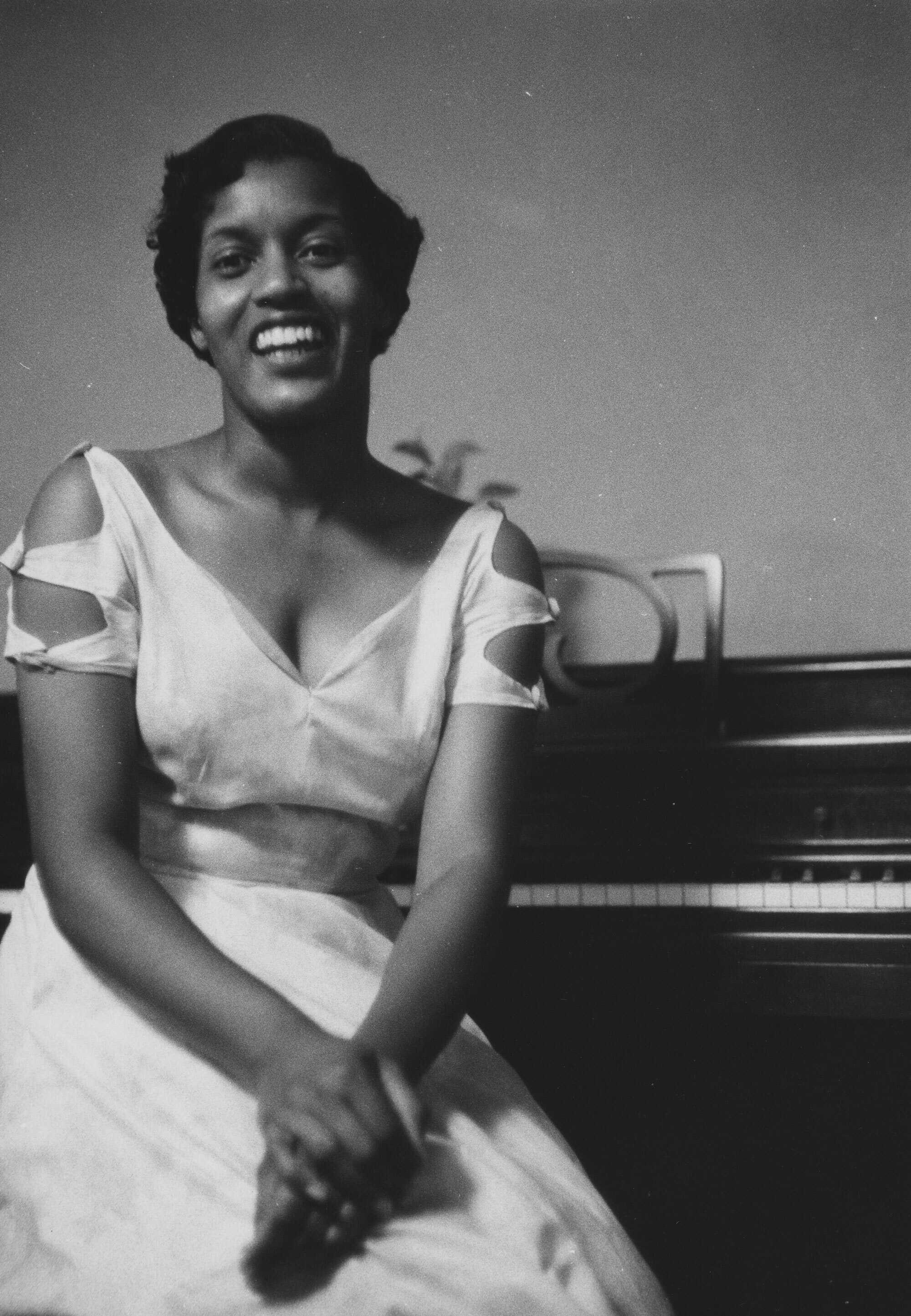 Black and white photograph of Myrlie Evers seated at a piano dressed in an elegant white dress.