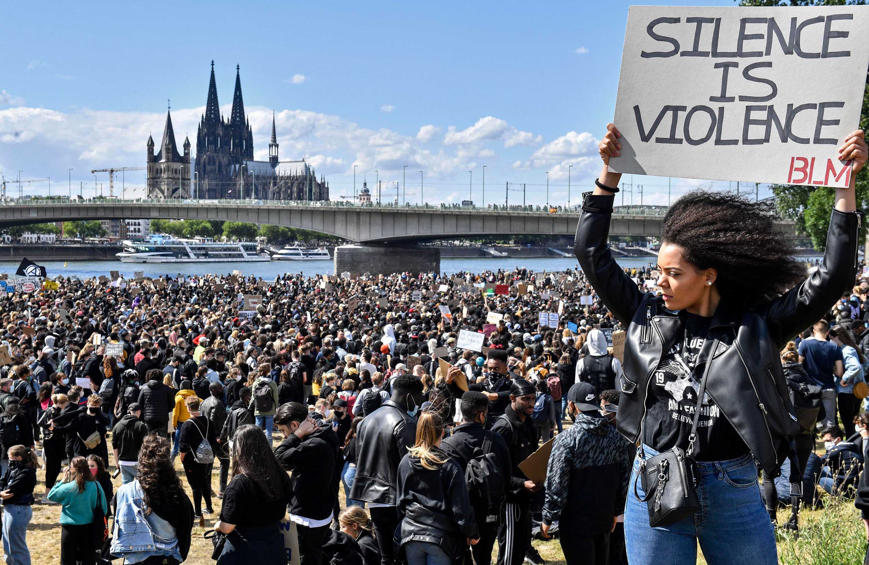 A Black Lives Matter protest in Cologne, Germany along the river. A Black woman is holding a protest sign in the foreground.