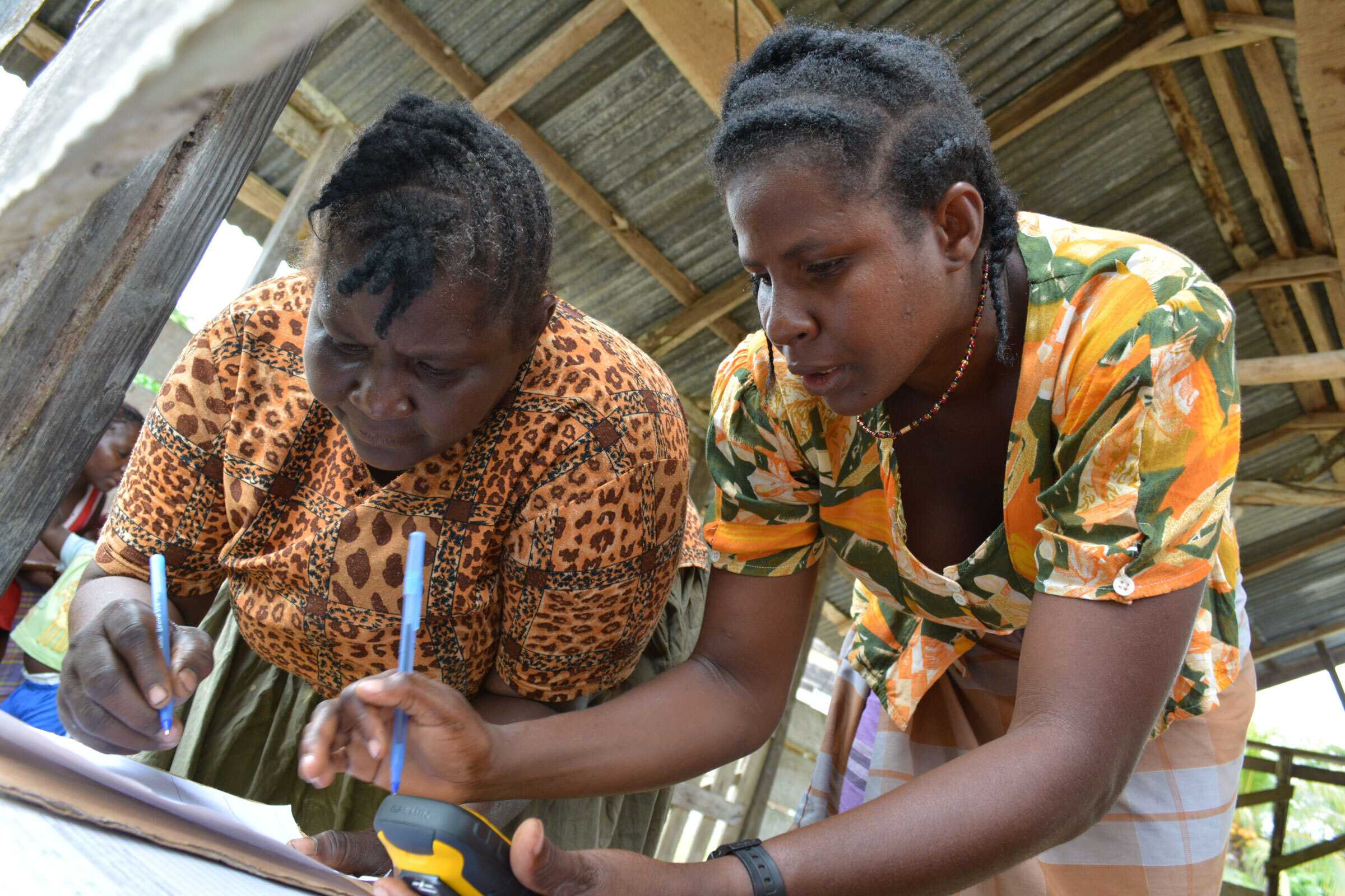 Two women sign papers on clipboards in an open market. They both are wearing yelllow patterned shirts.