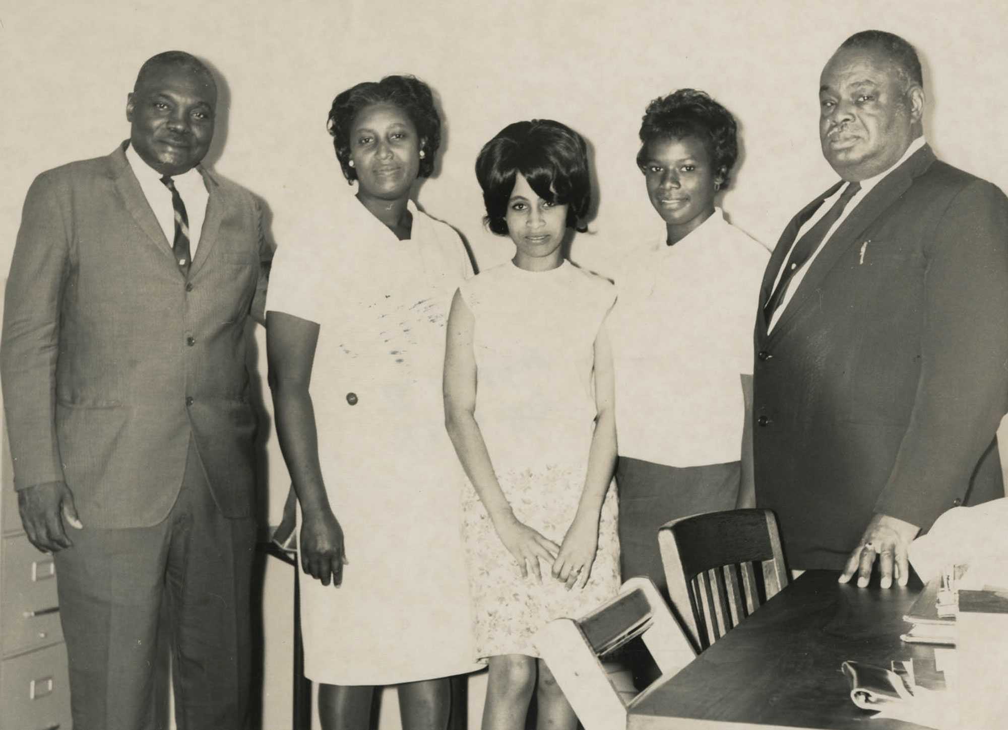 A black and white photograph of 5 members of the Community Owned Credit Union and Citizens Committee. They stand together in the office, next to a desk.