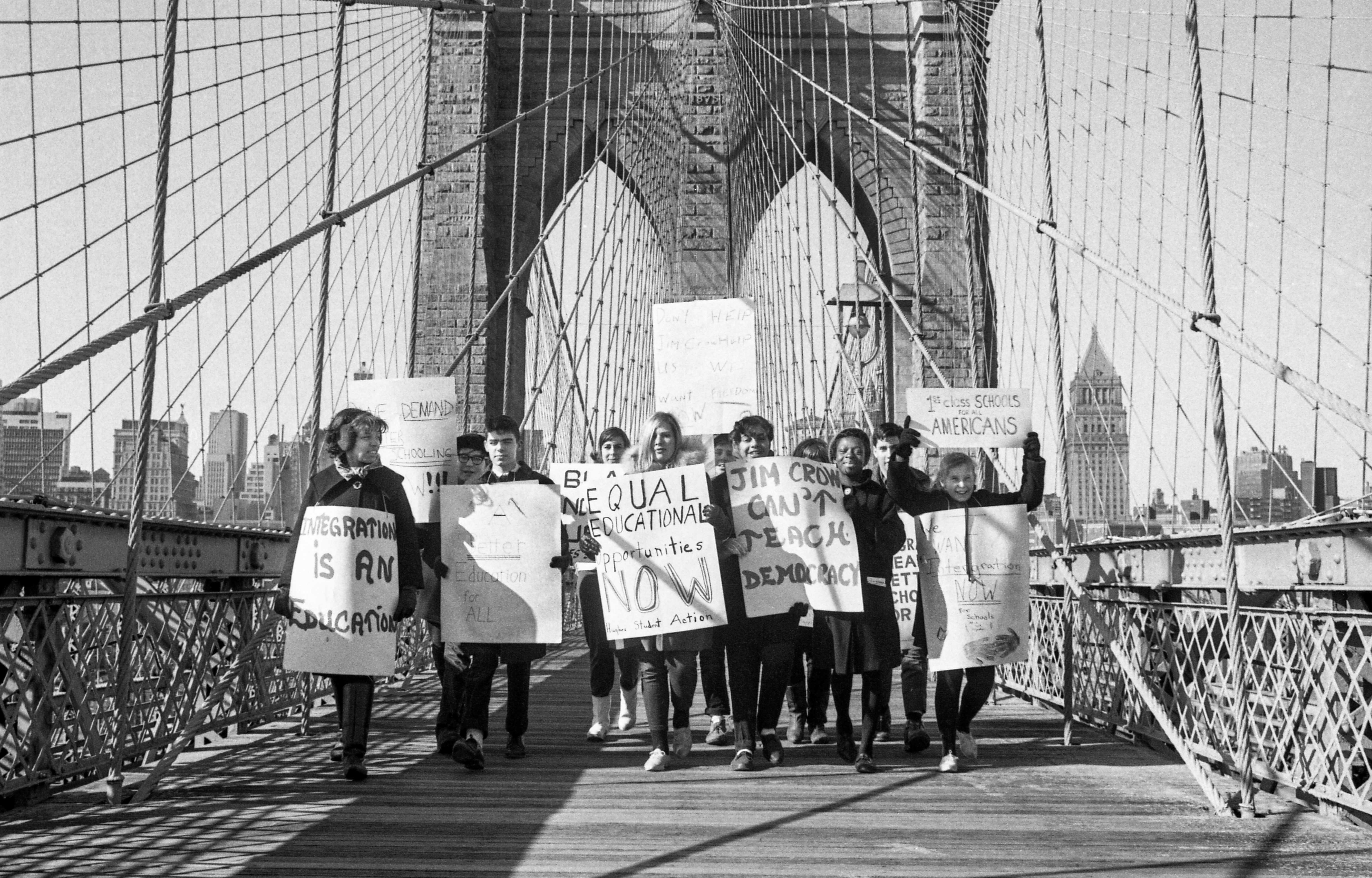 Black and white photograph of students and teachers standing on a bridge with protest signs decrying school segregation.