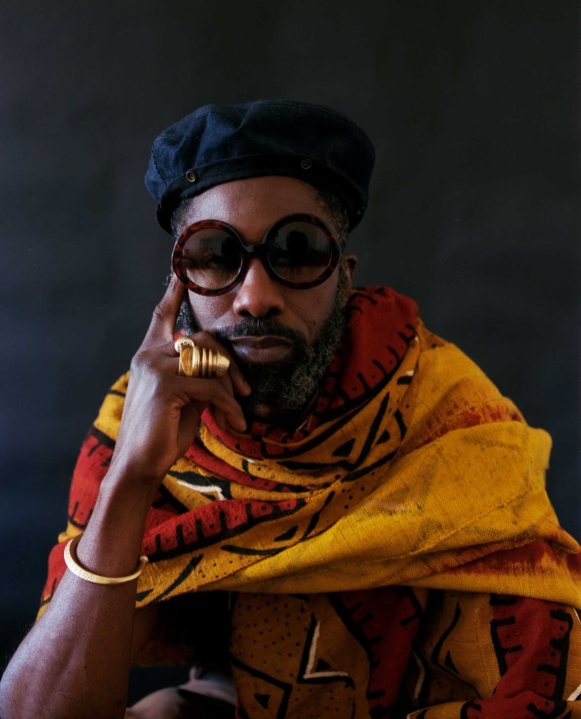 Artist Nyugen E. Smith poses for a portrait. He is wrapped in a gold fabric, large circular sunglasses, and dark hat.