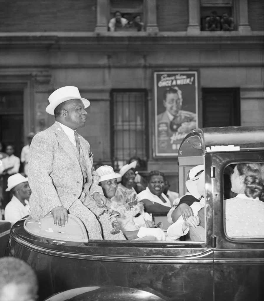 Black and white photo of an African American man seated in the back of a roofless vehicle.  He appears to be riding in a parade of some sort.  There are people crowded by the vehicle.