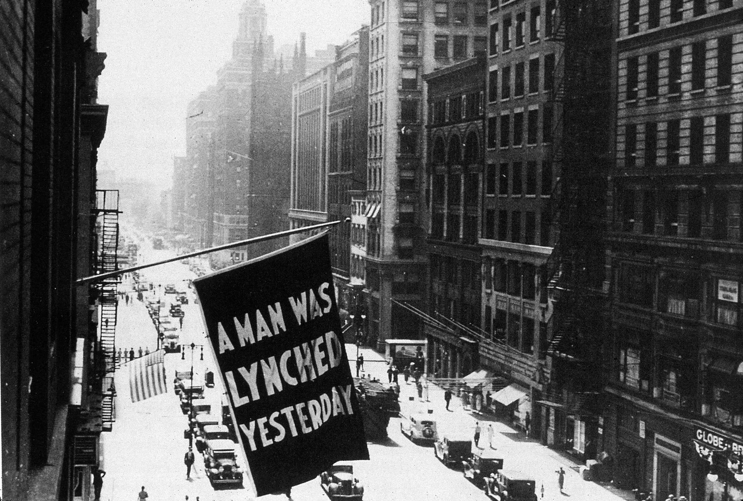 Black and white photograph of city buildings, one with a flag "A MAN WAS LYNCHED YESTERDAY" written on it.