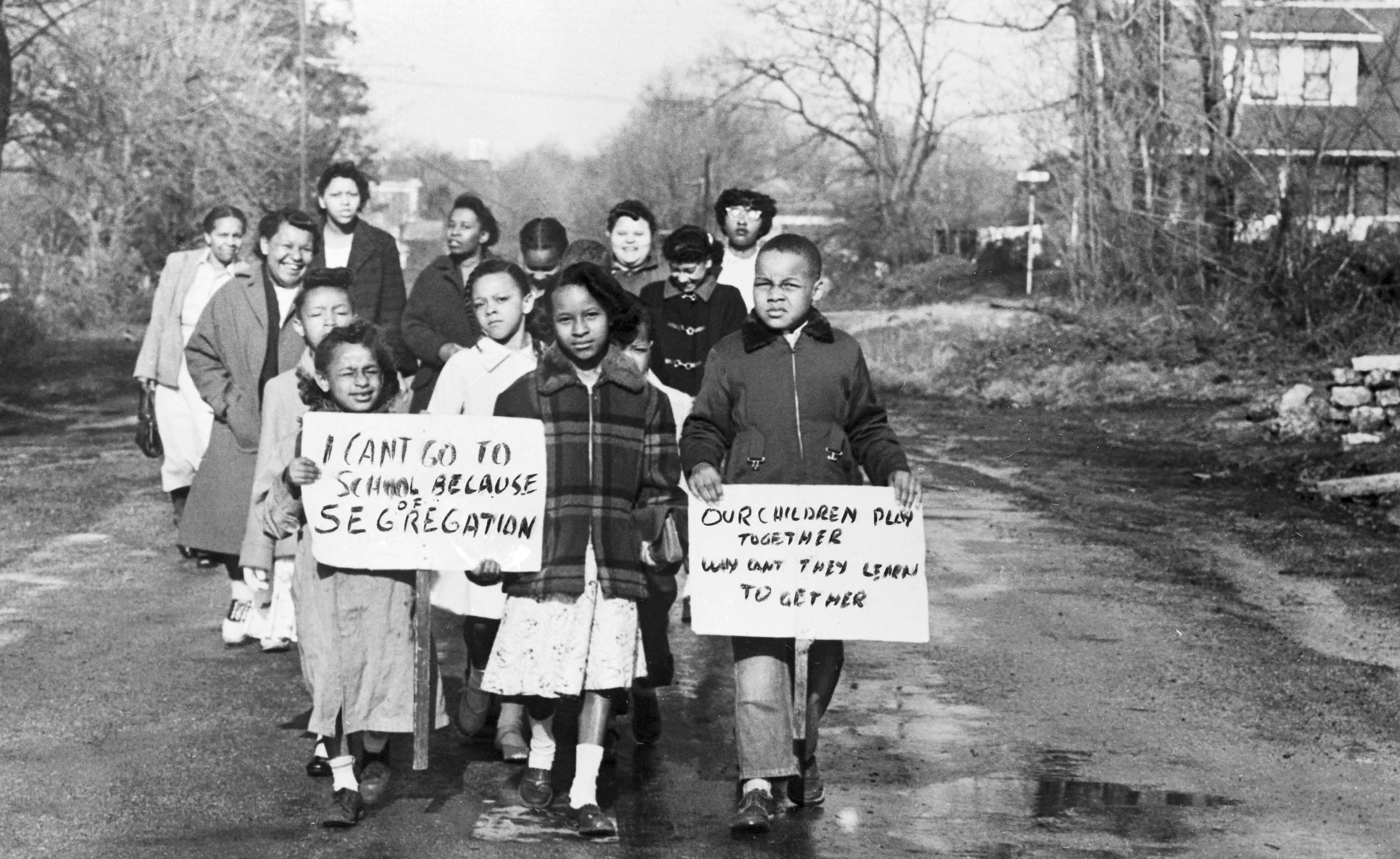 Black and white photograph of a group of African-American Children standing in a group protesting segregation.  Two of them are holding signs  "I Can't Go To Shcool Because of Segregation" and "Our Children Play Together Why Can't they Learn Together"
