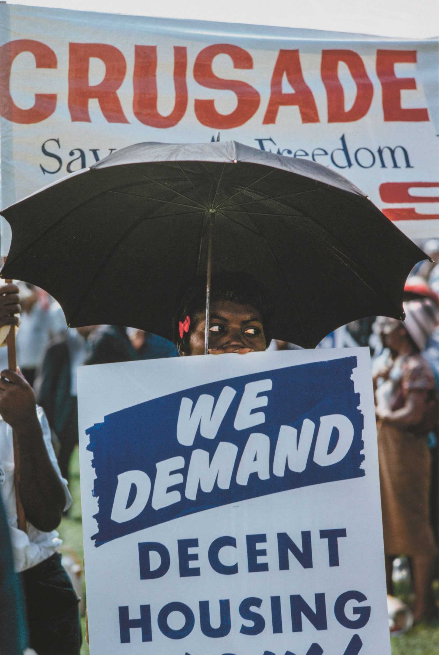 Color photograph from the 1963 March on Washington, showing a person holding a sign reading "We demand decent housing," with a black umbrella and pink bow in their hair.