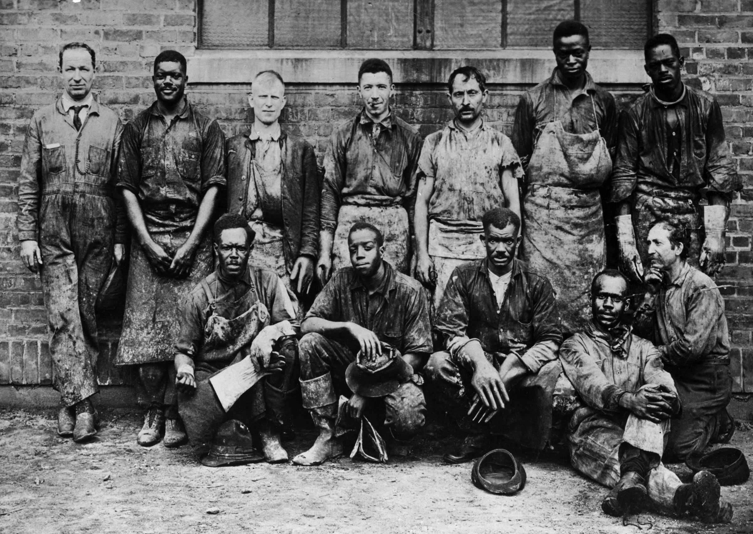 Black and white group photograph of men standing and sitting in front a brick building.  The men are dressed in workman's clothing.