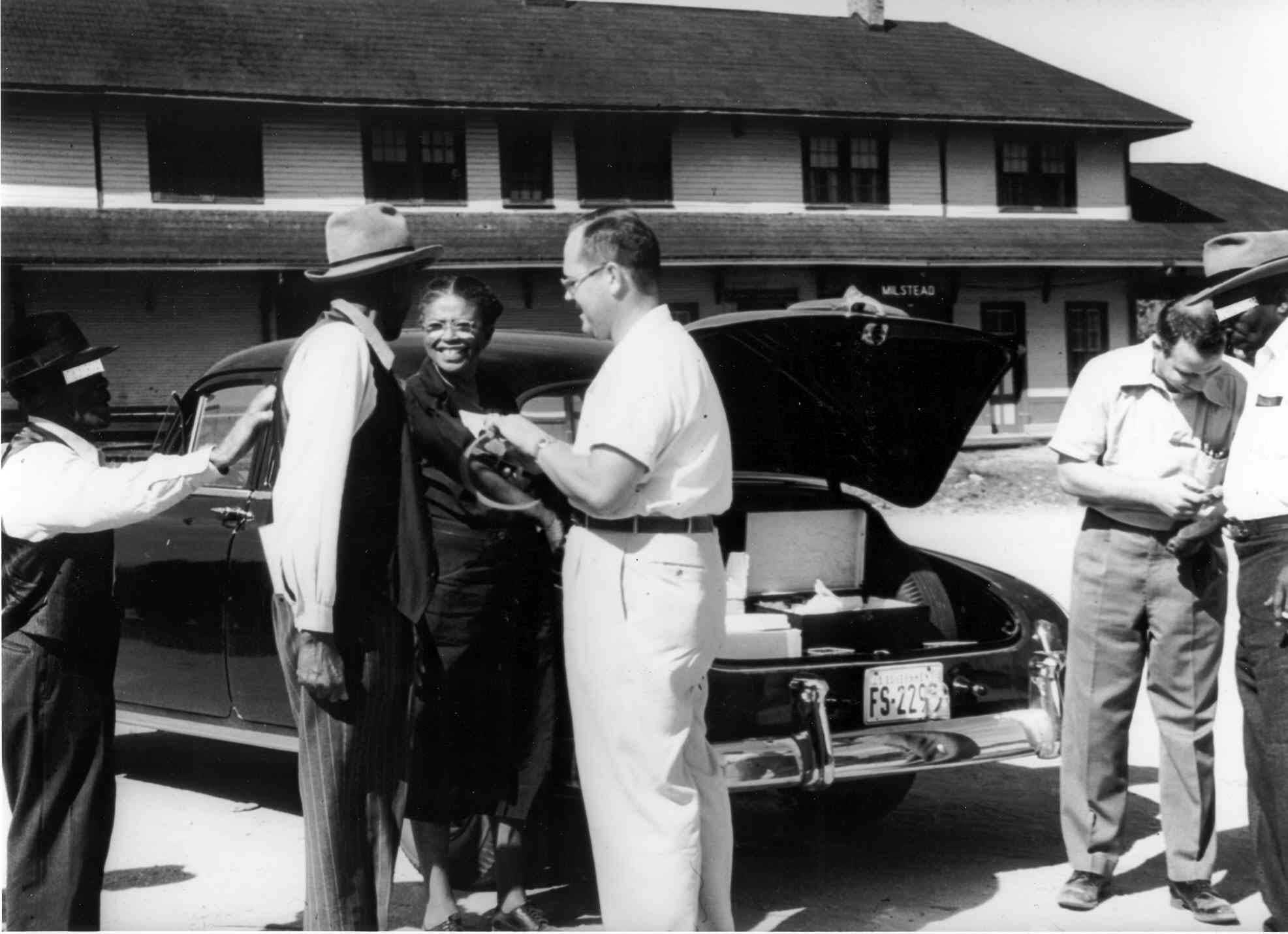 Black and white photograph showing white men interviewing African American people standing by a car with its trunk open.