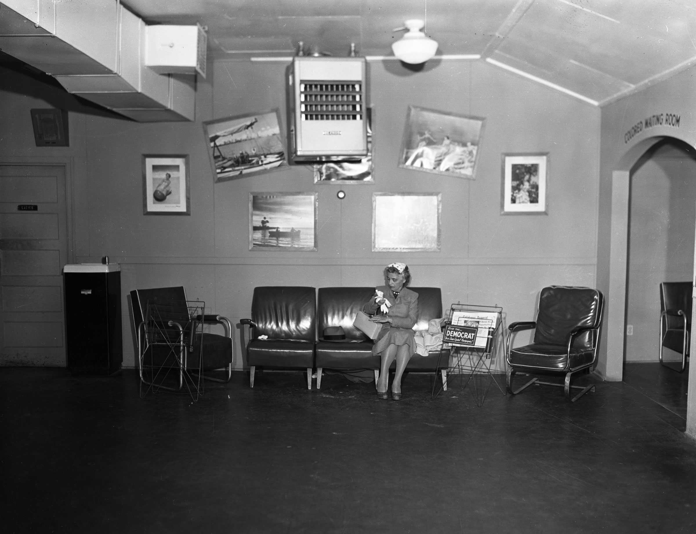 Black and white photograph showing interior of a waiting room.  There is a woman seated on a leather couch.  Behind her there are framed photographs and to her left there is a sign "COLORED WAITING ROOM" painted on the wall.