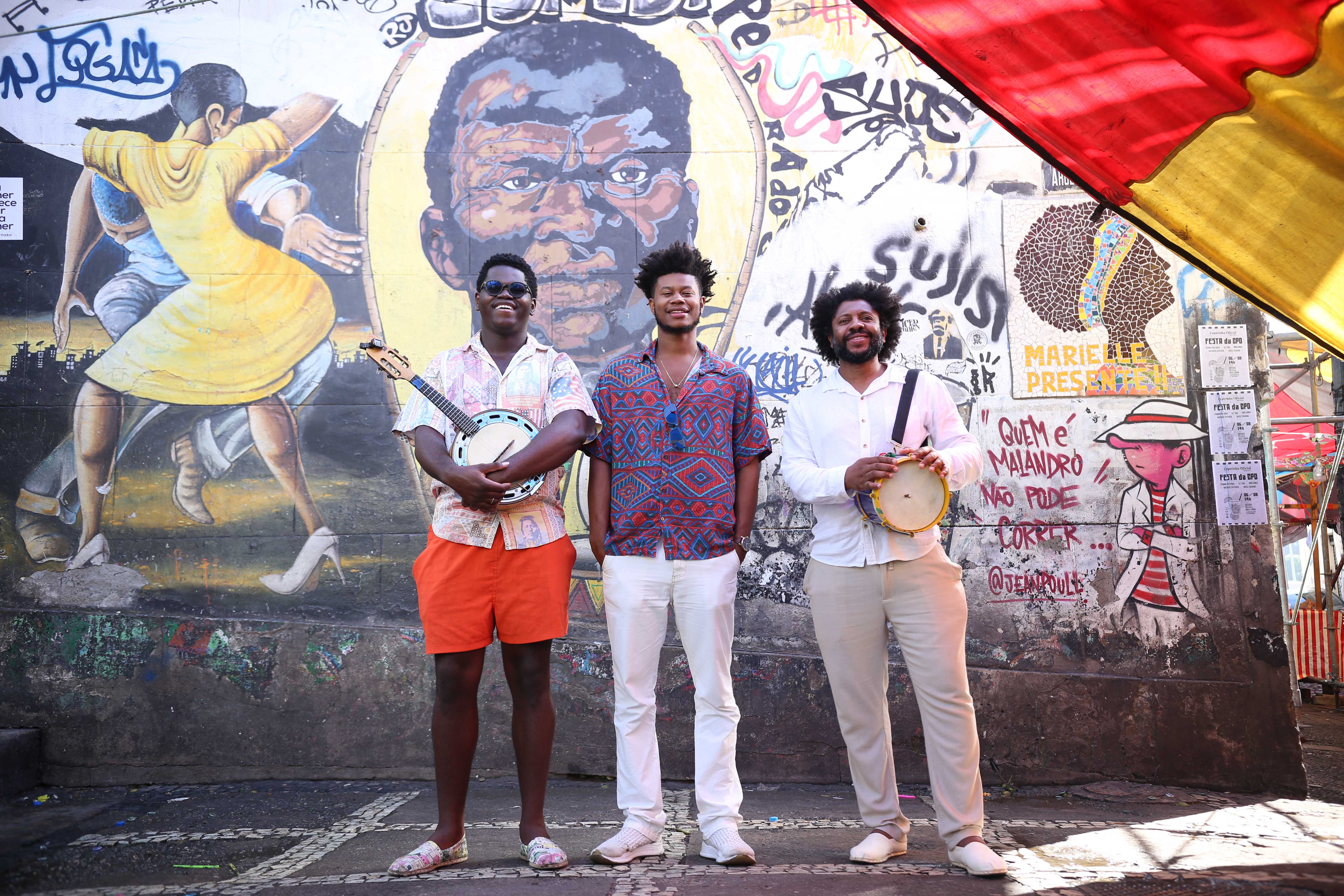 Alcino Amaral stand with two other men in front of a mural. The two men hold various instruments.